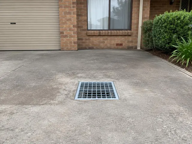 Concrete driveway with a square metal drain grate beside a brick house in Thornton